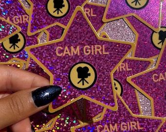 May include: A collection of star-shaped stickers with a glittery purple finish. Each sticker features the text "CAM GIRL" in gold lettering, along with a gold circle containing a black film camera icon. The stickers are being held by a hand.