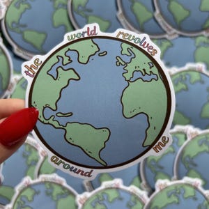 May include: A round sticker featuring a cartoon illustration of Earth with continents in green and oceans in blue. The text "the world revolves around me" is written around the globe in a rainbow font.