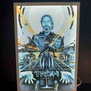 Peut inclure: Une affiche encadrée et rétro-éclairée du film "Tron: Legacy". L'affiche présente une image centrale d'un visage d'homme, avec d'autres personnages et éléments du film. L'affiche a une palette de couleurs bleue et jaune, avec le titre du film en bas.