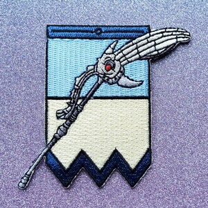 May include: An embroidered patch featuring a white skeletal hand holding a staff with a red gem. The staff is on a blue and white banner with a blue and white zig-zag border.