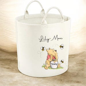 May include: Off-white felt storage basket with two handles. The basket features a Winnie the Pooh illustration with bees and the name "Lily-Mae" in script. The illustration shows Winnie the Pooh with a honey pot.