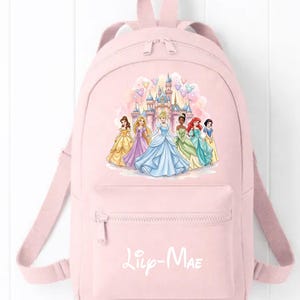 May include: A light pink backpack featuring a graphic of seven Disney princesses standing in front of a castle with colourful balloons. The front pocket displays the name Lily-Mae in white script lettering. The bag has adjustable shoulder straps.