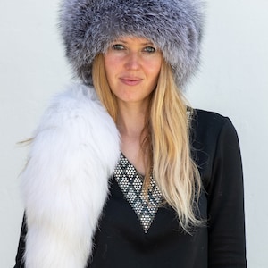 May include: A woman wearing a grey and white fur hat and a white fur scarf. She is wearing a black shirt with a sparkly neckline.