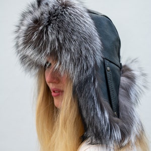 May include: A black leather and silver fox fur hat with ear flaps. The hat is worn by a person with long blonde hair.