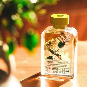 May include: A clear glass bottle with a yellow lid filled with a liquid and a small bouquet of white flowers and green leaves. The label reads "Jazmine x Le Petit Jardin & Co." and "The uplifting fresh morning aromas of blooming Jasmine in the Amazon rainforest". The bottle also has a label that reads "200ml 60", "6.803", "DIFFUSER", and "#4".