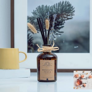 May include: A brown glass diffuser bottle with a label that reads "Le Petit Jardin & Co. Moscato Pesca Peach Moscato" and a natural fiber bow. The bottle is filled with a clear liquid and has black diffuser sticks inserted. The bottle is surrounded by dried lavender and pampas grass.
