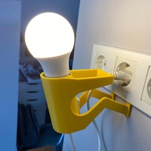 Può includere: Un supporto da parete giallo stampato in 3D con una lampadina bianca collegata a una presa a muro. Il supporto ha la forma di una persona che tiene la lampadina.