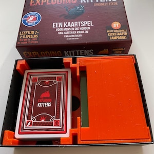 Peut inclure: Boîte de jeu de cartes Exploding Kittens ouverte. Le couvercle de la boîte présente le titre du jeu et une illustration de chat de dessin animé. À l'intérieur, un jeu de cartes avec un design rouge et blanc est visible, ainsi que des composants de jeu orange.