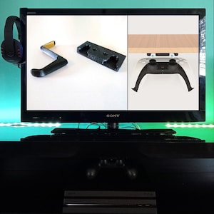 Op de afbeelding: Een zwarte tv met een wit scherm waarop twee afbeeldingen te zien zijn van een zwarte en gele controllerhouder en een witte controllerhouder. De tv staat op een zwarte standaard met een zwart en zilver apparaat eronder.