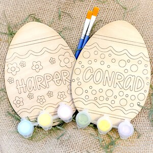 May include: Two unfinished wooden Easter egg shapes with the names "Harper" and "Conrad" written on them. The eggs are surrounded by paint pots and paintbrushes.