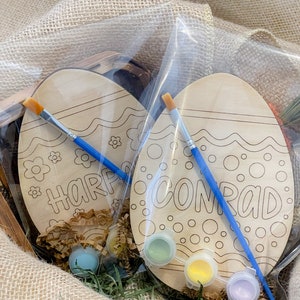 May include: Two wooden Easter eggs with paint and brushes in a burlap bag. The eggs are decorated with a wavy line and dots. One egg says "Harper" and the other says "Conrad".