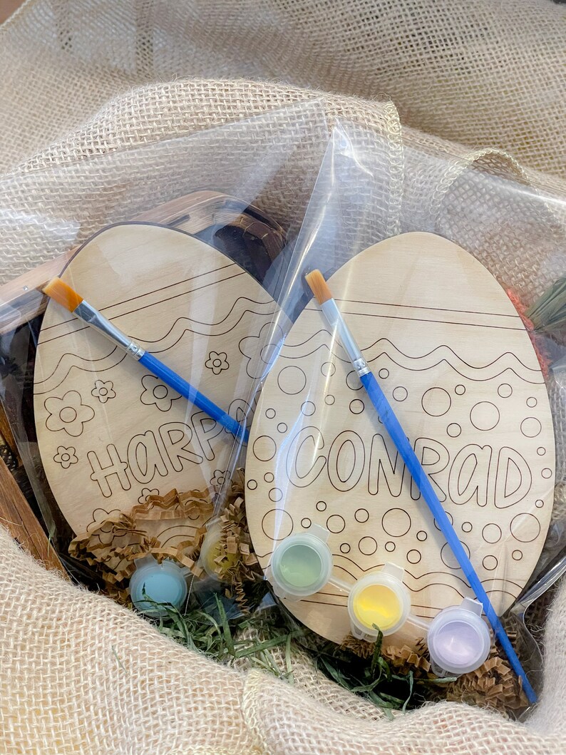 May include: Two wooden Easter eggs with paint and brushes in a burlap bag. The eggs are decorated with a wavy line and dots. One egg says "Harper" and the other says "Conrad".