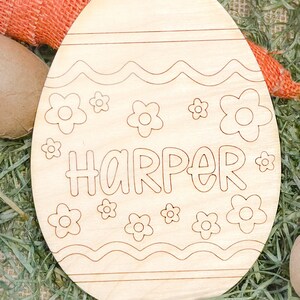 May include: A wooden Easter egg shaped ornament with a wavy line design and flowers. The name "HARPER" is written in a whimsical font on the egg.