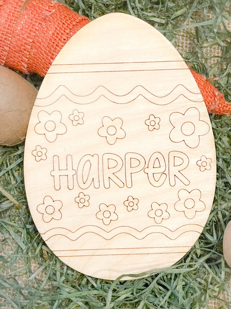 May include: A wooden Easter egg shaped ornament with a wavy line design and flowers. The name "HARPER" is written in a whimsical font on the egg.