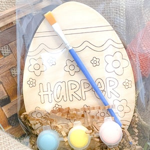 May include: A wooden Easter egg shaped craft kit with the name "Harper" written on it. The kit includes paint, a paintbrush, and brown paper shreds.