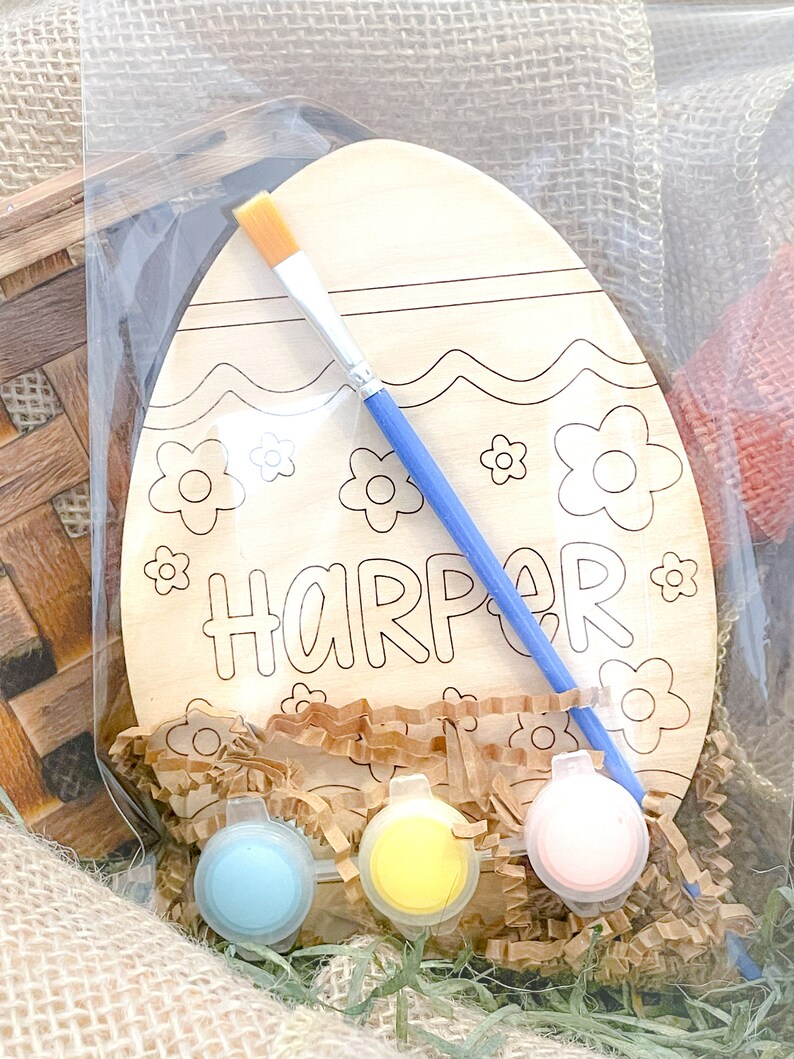 May include: A wooden Easter egg shaped craft kit with the name "Harper" written on it. The kit includes paint, a paintbrush, and brown paper shreds.