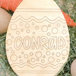 May include: A wooden Easter egg shaped ornament with a white outline of a wavy design and polka dots. The name "CONRAD" is written in a white outline on the egg.