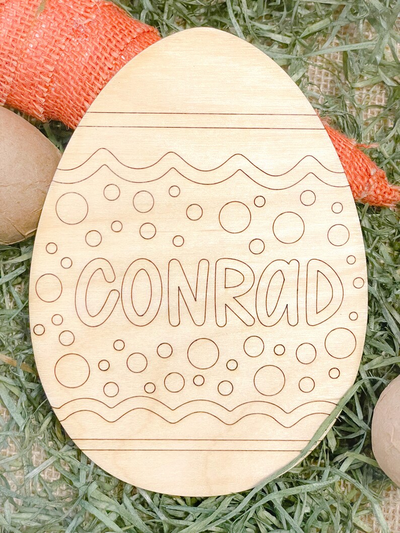 May include: A wooden Easter egg shaped ornament with a white outline of a wavy design and polka dots. The name "CONRAD" is written in a white outline on the egg.