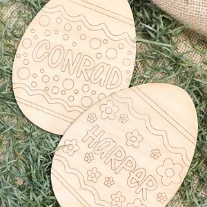 May include: Two unfinished wooden Easter eggs with the names Conrad and Harper written on them. The eggs are decorated with a wavy line pattern and small circles and flowers.