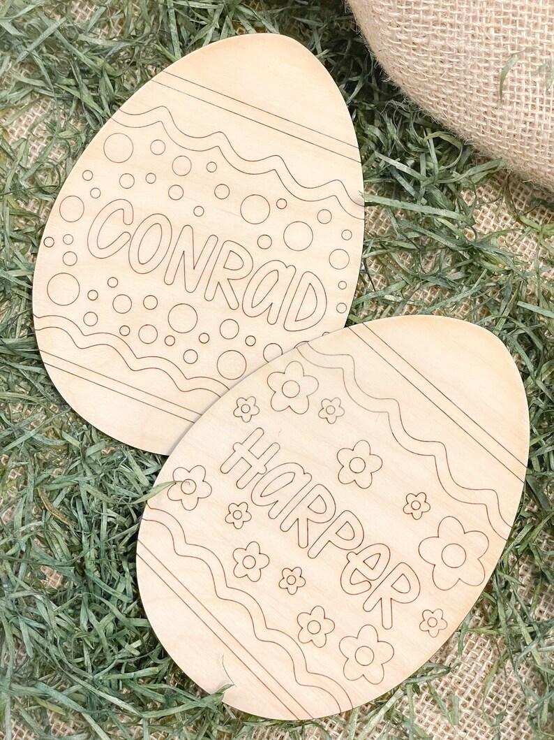May include: Two unfinished wooden Easter eggs with the names Conrad and Harper written on them. The eggs are decorated with a wavy line pattern and small circles and flowers.