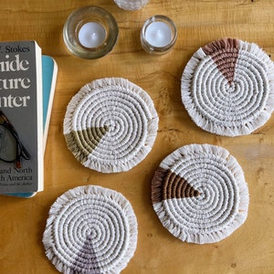 May include: Four round crocheted coasters with fringe edges. Each coaster has a different color accent in the center: brown, green, purple, and brown. The coasters are on a wooden surface.