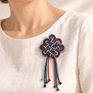 Könnte beinhalten: Eine dekorative Brosche mit einem komplexen geflochtenen Knotenmuster aus roter, grüner, blauer und silberner Kordel. Der mittlere Knoten ist wie eine stilisierte Blume geformt und hat sechs herabhängende Kordelstränge in passenden Farben.