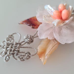 May include: A pink and white flower-shaped pendant with a silver knot and a brown and white shell. The pendant is made of carved stone and has a delicate design.