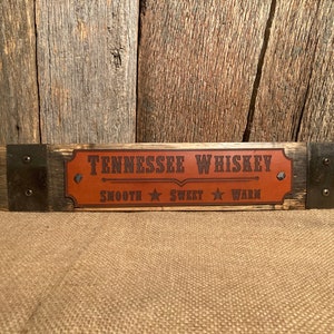 May include: A wooden sign with metal accents, featuring a brown leather patch with the text "Tennessee Whiskey" and the words "Smooth", "Sweet", and "Warm" printed below.