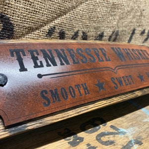 May include: A brown leather sign with black text that reads "Tennessee Whiskey Smooth Sweet Warm" with a star on either side of the words "Smooth" and "Sweet". The sign is attached to a wooden barrel.