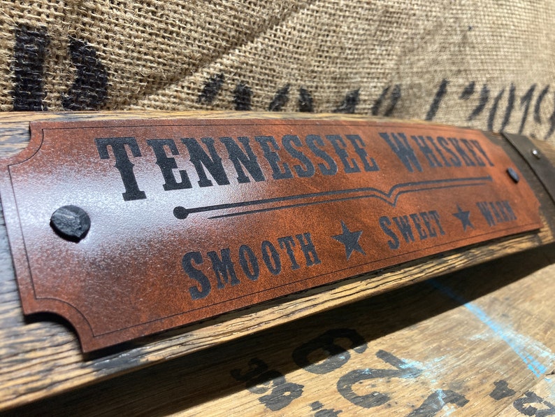May include: A brown leather sign with black text that reads "Tennessee Whiskey Smooth Sweet Warm" with a star on either side of the words "Smooth" and "Sweet". The sign is attached to a wooden barrel.