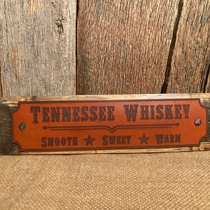 May include: A wooden sign with a brown leather patch that reads "Tennessee Whiskey" with a line underneath and the words "Smooth", "Sweet", and "Warm" with a star between each word.