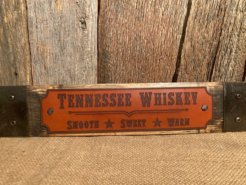 May include: A wooden sign with a brown leather patch that reads "Tennessee Whiskey" with a line underneath and the words "Smooth", "Sweet", and "Warm" with a star between each word.