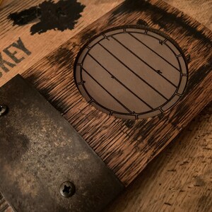 May include: A rustic wooden coaster with a barrel design etched in the center. The coaster is made from reclaimed wood and features a metal accent secured with screws. The wood has a dark, aged appearance, suggesting a whiskey barrel theme.