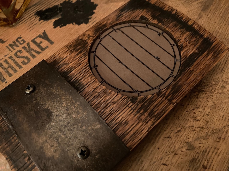 May include: A rustic wooden coaster with a barrel design etched in the center. The coaster is made from reclaimed wood and features a metal accent secured with screws. The wood has a dark, aged appearance, suggesting a whiskey barrel theme.