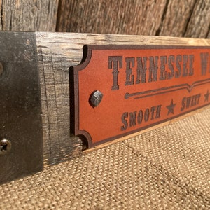 May include: A wooden sign with a brown leather plaque that reads "Tennessee Whiskey Smooth Sweet Wild". The sign is attached to a metal bracket with screws.