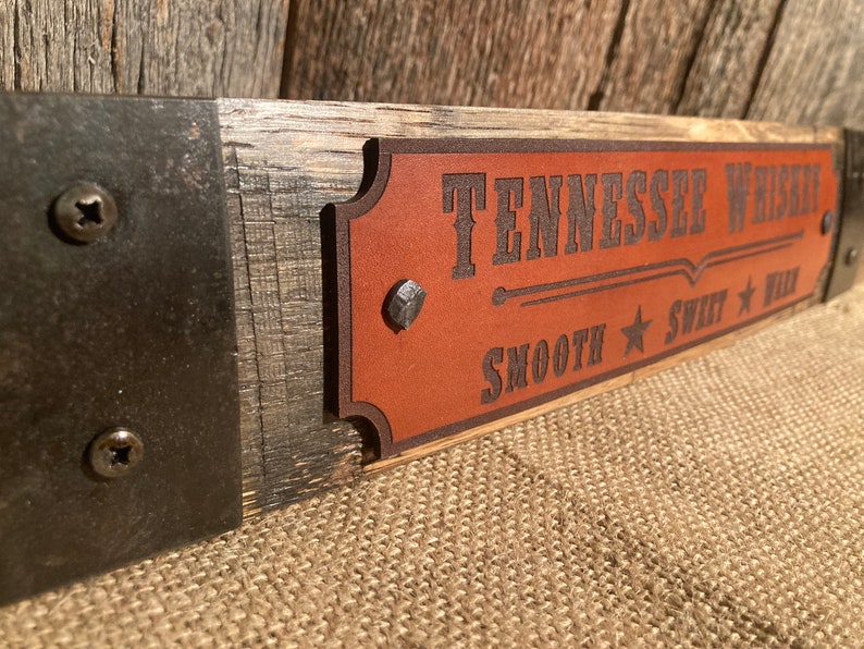 May include: A wooden sign with a brown leather plaque that reads "Tennessee Whiskey Smooth Sweet Wild". The sign is attached to a metal bracket with screws.