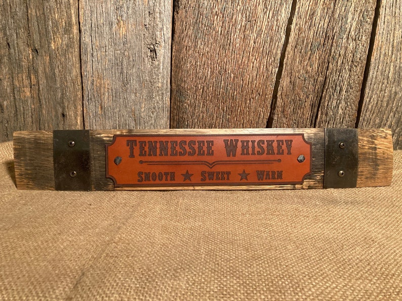May include: A wooden sign with metal accents, featuring a brown leather patch with the text "Tennessee Whiskey" and the words "Smooth", "Sweet", and "Warm" printed below.