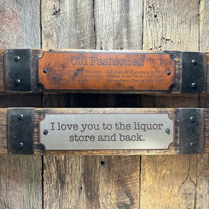 May include: Two wooden signs with metal accents hanging on a wood wall. The top sign has a brown leather plaque with the text "Old Fashioned" and the recipe for the cocktail. The bottom sign has a silver plaque with the text "I love you to the liquor store and back."
