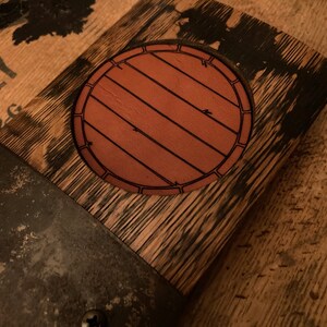 May include: Close-up of a wooden barrel head with a circular, brown leather emblem featuring a barrel design. The wood has a dark, weathered appearance, with metal bands and screws. The overall theme is rustic and vintage.