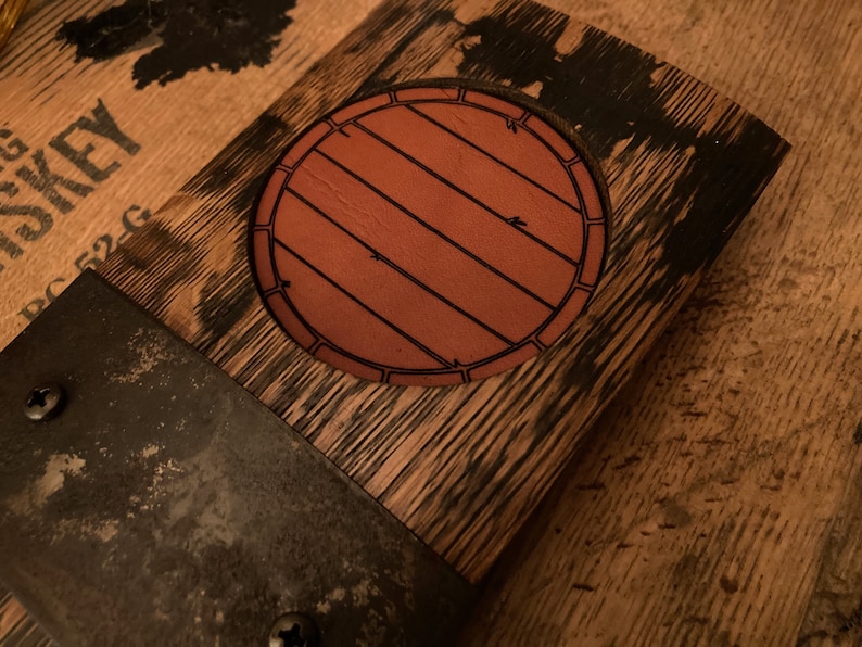 May include: Close-up of a wooden barrel head with a circular, brown leather emblem featuring a barrel design. The wood has a dark, weathered appearance, with metal bands and screws. The overall theme is rustic and vintage.