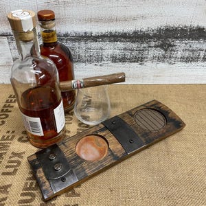 May include: A wooden cigar cutter with a round opening and a metal blade, next to a glass, a lit cigar, and two bottles of liquor. The bottles are filled with a dark amber liquid.