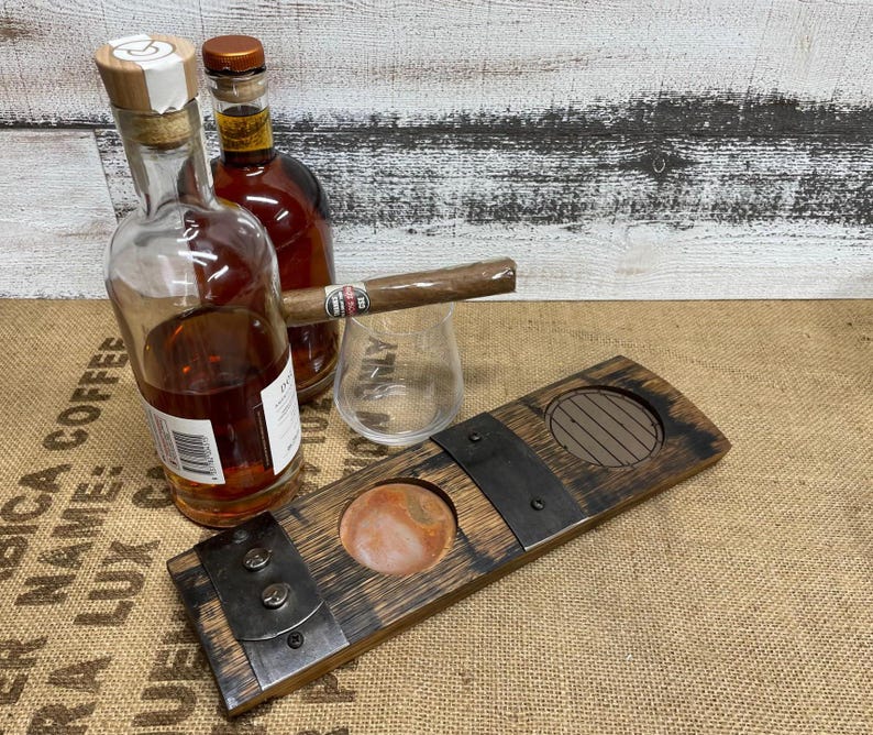 May include: A wooden cigar cutter with a round opening and a metal blade, next to a glass, a lit cigar, and two bottles of liquor. The bottles are filled with a dark amber liquid.