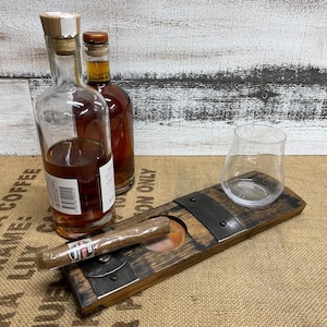 May include: A wooden cigar ashtray with a built-in cigar rest and a clear glass. Two bottles of amber-colored liquid are in the background. The ashtray is made of dark wood and metal accents.