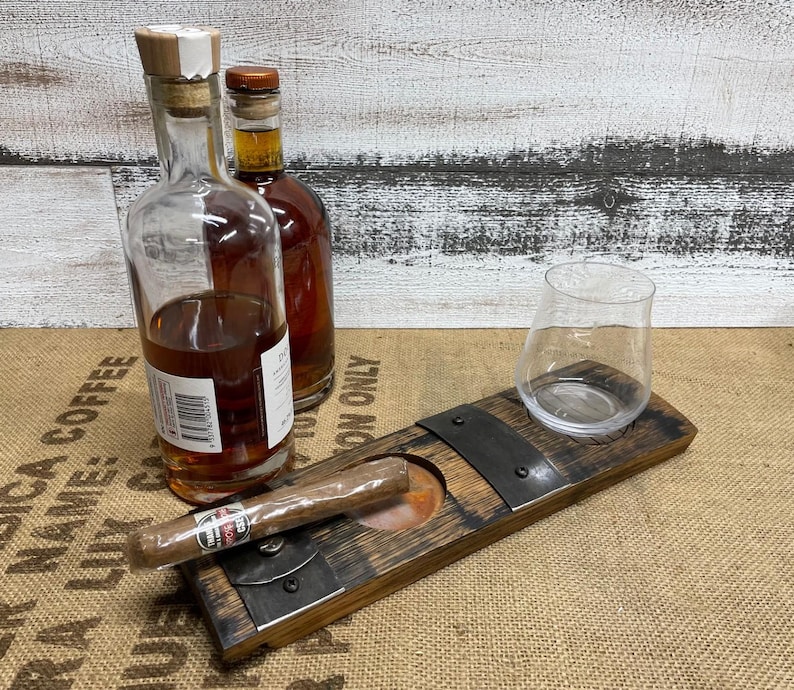 May include: A wooden cigar ashtray with a built-in cigar rest and a clear glass. Two bottles of amber-colored liquid are in the background. The ashtray is made of dark wood and metal accents.