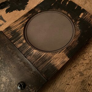 May include: A rustic coaster made from a section of a whiskey barrel. The coaster has a dark brown, circular inset for a glass, and is accented with metal hardware and screws. The wood grain is visible, and the edges are charred.