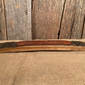 May include: A wooden sign made from a reclaimed whiskey barrel stave with a leather label that reads "Tennessee Whiskey Smooth Sweet Warm". The sign has two metal brackets with rivets on each end.