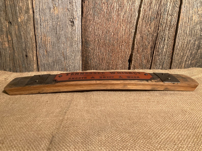 May include: A wooden sign made from a reclaimed whiskey barrel stave with a leather label that reads "Tennessee Whiskey Smooth Sweet Warm". The sign has two metal brackets with rivets on each end.