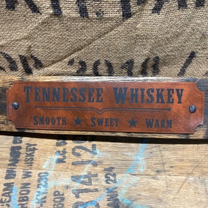 May include: A wooden sign with a brown leather plaque that reads "Tennessee Whiskey" with the words "Smooth", "Sweet", and "Warm" underneath. The sign is made from a reclaimed whiskey barrel stave.