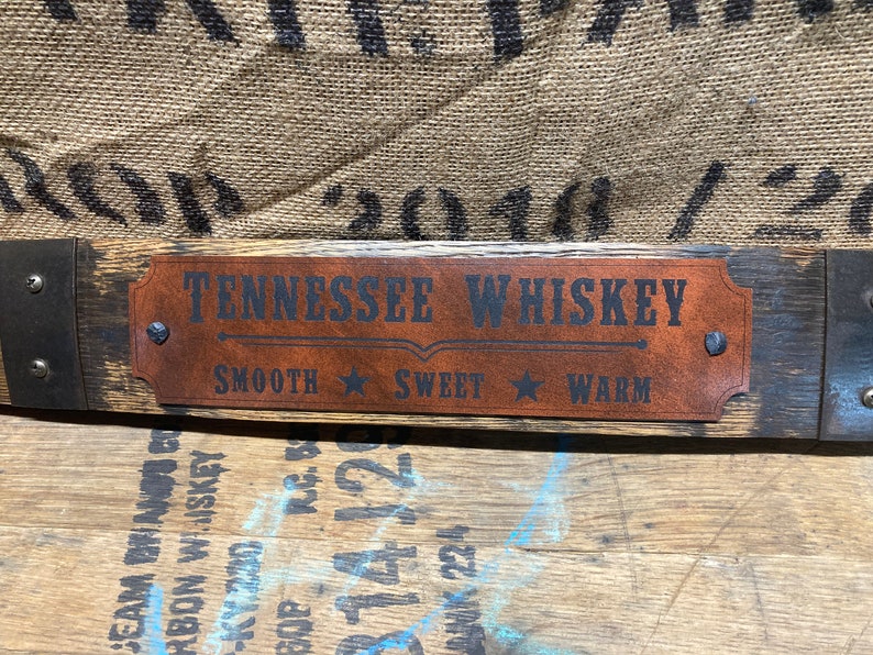 May include: A wooden sign with a brown leather plaque that reads "Tennessee Whiskey" with the words "Smooth", "Sweet", and "Warm" underneath. The sign is made from a reclaimed whiskey barrel stave.
