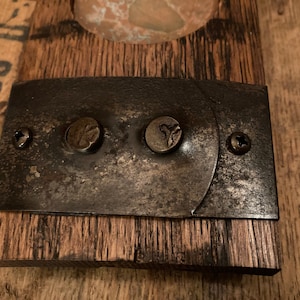 May include: Close-up of a rustic metal plate with two rivets and two screws, attached to a piece of weathered wood. The metal has a distressed, aged appearance. The wood has a natural, rough texture.
